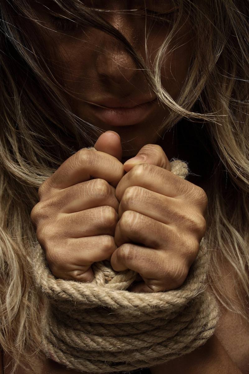 Mulher de cabeça baixa com as mãos amarradas por uma corda, simbolizando a violência que prende a vítima.